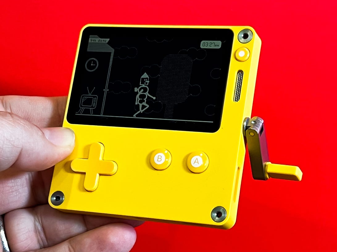 A hand holding the bright yellow Panic Playdate gaming device in front of a vivd red background.